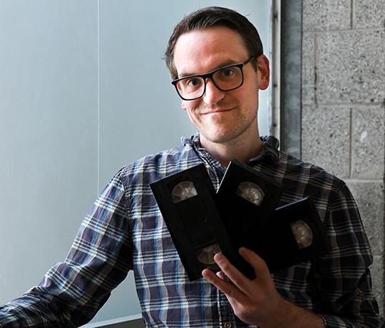 man with glasses holding VHS tapes and grinning